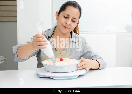 Donna ispanica nella sua pasticceria che decora una torta - donna mettere la panna montata sulla torta appena sfornata - donna intraprendente Foto Stock