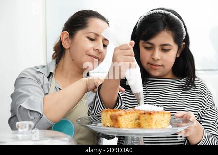 Mamma e figlia ispaniche che decorano una torta con panna montata - mamma latina insegna a sua figlia di decorare una torta Foto Stock