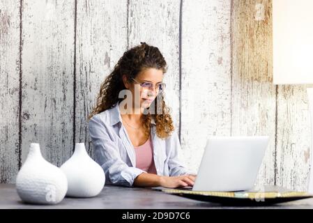 Giovane donna che indossa bicchieri seduti a un tavolo da pranzo remoto lavorare su un computer portatile da casa durante il blocco per La pandemia di Covid-19 Foto Stock