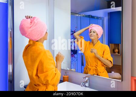 Giovane donna che indossa un accappatoio arancione dai colori vivaci con lei capelli in un asciugamano dopo la doccia guardando se stessa in uno specchio da bagno con un s tranquillo Foto Stock