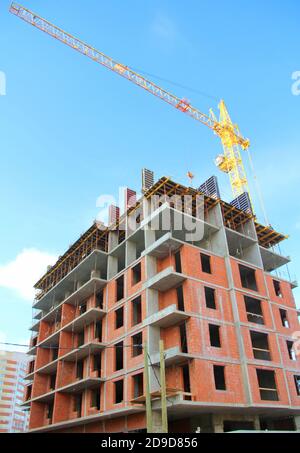 Un edificio di appartamenti a più piani e' in costruzione con mattoni e cemento. Costruzione dell'edificio contro il cielo blu. La gru a torre è gialla. Foto Stock