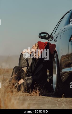 Elegante donna brunette in caldo cappotto grigio e lavorato a maglia cappello Bordeaux è seduto a terra vicino alla sua auto. Auto-viaggio locale, immagine colorata stile di vita Foto Stock