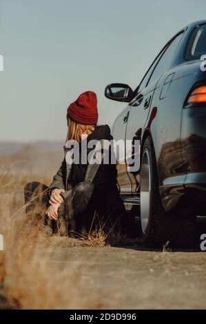 Donna elegante senza viso in caldo cappotto grigio e lavorato a maglia cappello Bordeaux è seduto a terra vicino alla sua auto. Auto-viaggio locale, immagine colorata stile di vita Foto Stock