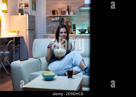 Donna felice che mangia popcorn sul divano e guarda la tv nel soggiorno di casa. Eccitata, divertita, solitaria, che si gode la serata seduta su un comodo divano vestito in pigiama davanti alla televisione. Foto Stock