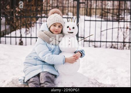 Ragazza e pupazzo di neve. Una ragazza adolescente carina in un cappello di maglia invernale e una giacca blu giù con il suo amico lo pupazzo di neve. Foto Stock