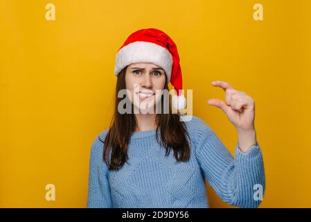 Giovane donna mostra oggetto molto minuscolo, forma qualcosa di piccolo, indossa cappello di Natale e maglione blu, sguardi femminile in puzzle, bisogno di un po 'di più Foto Stock