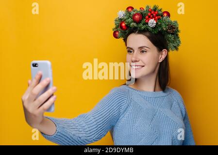 Felice giovane donna con un sorriso piacevole tenere i telefoni in mani rende selfie, indossa abete rosso corona di vacanza intorno alla testa, fa videochiamata Foto Stock