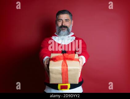 Ritratto di un uomo bearded in felpa di natale che dà dono box e guardando la telecamera isolata su sfondo rosso Foto Stock