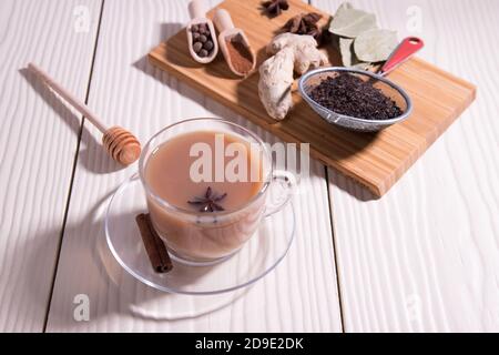 Una tazza di tè caldo indiano masala tè, cucchiai con spezie, zenzero, anice su un tagliere, su uno sfondo bianco. Orientamento orizzontale. Foto Stock