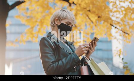 Giovane donna con maschera facciale e borse per la spesa utilizzando smartphone sotto l'albero con foglie gialle. Foto di alta qualità Foto Stock