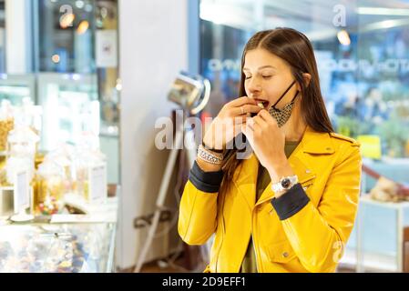 Donna giovane e felice che indossa una maschera protettiva per il viso che mangia un cioccolato bianco in piedi all'interno di un shop.girl avendo una caramella dolce in autunno dopo il blocco Foto Stock