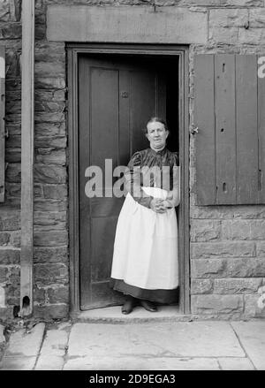 Una signora anziana che indossava un vestito a maniche e un lungo grembiule si trovava all'ingresso di una casa. Immagine di epoca vittoriana, circa 1900 in bianco e nero. Scansionato dal negativo originale e ripristinato. Foto Stock