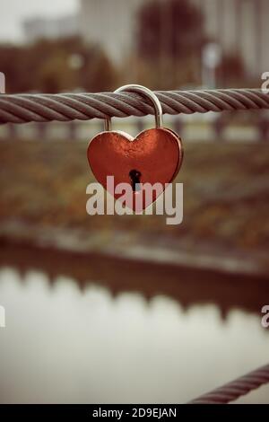 Lucchetto in metallo rosso a cuore su cavo in acciaio della recinzione del ponte. Paesaggio urbano non focalizzato sullo sfondo. Orientamento verticale. Messa a fuoco selettiva. Spazio di copia. Matrimonio Foto Stock