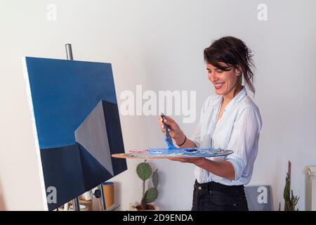 Un giovane pittore sorridente mescola colori su una tavolozza nel suo studio di casa. Dietro di esso, si può vedere un'opera d'arte contemporanea messa su un cavalletto, così come Foto Stock