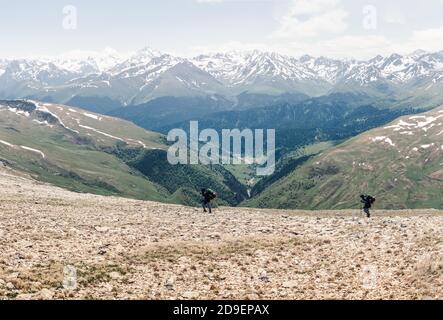 Vista dall'alto due viaggiatori arrampicatori con zaini e bastoni salire alta montagna che domina la valle verde e cime innevate del montagne contro Foto Stock