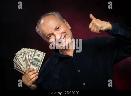 Signore anziano che tiene una pila di soldi. Ritratto di un vecchio imprenditore eccitato. Felice vecchio uomo che tiene le banconote del dollaro su sfondo rosso scuro. Foto Stock