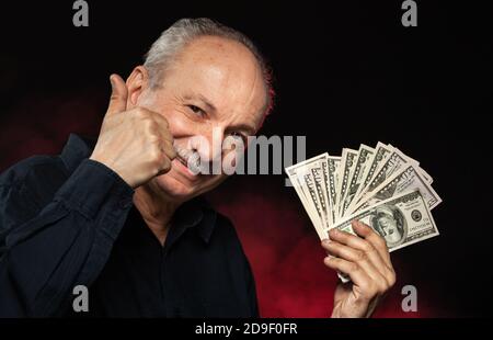 Signore anziano che tiene una pila di soldi. Ritratto di un vecchio imprenditore eccitato. Felice vecchio uomo che tiene le banconote del dollaro su sfondo rosso scuro. Foto Stock