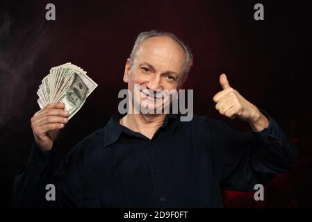 Signore anziano che tiene una pila di soldi. Ritratto di un vecchio imprenditore eccitato. Felice vecchio uomo che tiene le banconote del dollaro su sfondo rosso scuro. Foto Stock