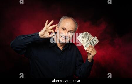 Signore anziano che tiene una pila di soldi. Ritratto di un vecchio imprenditore eccitato. Felice vecchio uomo che tiene le banconote del dollaro su sfondo rosso scuro. Foto Stock