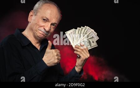Signore anziano che tiene una pila di soldi. Ritratto di un vecchio imprenditore eccitato. Felice vecchio uomo che tiene le banconote del dollaro su sfondo rosso scuro. Foto Stock