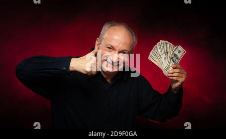 Signore anziano che tiene una pila di soldi. Ritratto di un vecchio imprenditore eccitato. Felice vecchio uomo che tiene le banconote del dollaro su sfondo rosso scuro. Foto Stock