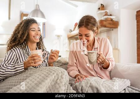Abitudini familiari con diversità: Coppia omosessuale multirazziale femmina divertirsi a casa seduta sul divano bere una tazza di tè sotto una coperta - due Foto Stock
