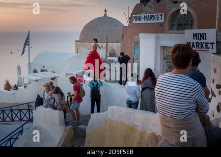 Una sposa orientale nel suo vestito rosso brillante con il suo sposo in presenza ha le sue foto scattate su un tetto piatto come turisti si riuniscono per guardare il famoso tramonto nella città di Oia sull'isola greca di vacanze di Santorini. 19 ottobre 2015. Foto: Neil Turner Foto Stock