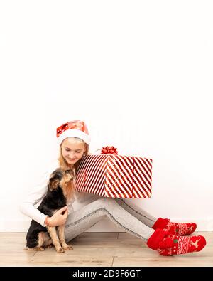 Una bella bambina in abiti casual con accessori di Natale si siede sul pavimento e abbraccia una grande scatola regalo con un nastro rosso e un arco. Lunghezza intera Foto Stock