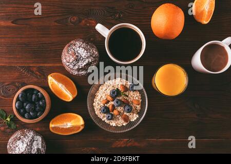 Composizione piatta di cibi sani per la colazione con farinata d'avena, frutta secca, frutti di bosco, succo d'arancia, muffin e caffè fresco su sfondo di legno. Foto Stock
