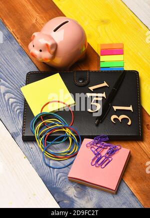 Composizione Home Finance. Banca piggy e cancelleria su sfondo colorato di legno. Moneybox con viso di maiale da organizer, note appiccicose, segnalibri, penna, elastici e numeri. Concetto di investimento e denaro Foto Stock