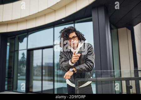Bell'uomo giovane con capelli neri ricci e in occhiali si trova sulla strada contro l'edificio Foto Stock