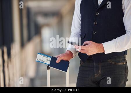 Vista ravvicinata del passeggero maschile in abiti eleganti e formali si trova nella sala dell'aeroporto con bagagli e biglietti Foto Stock
