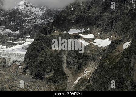 Nel mese di agosto, mesi prima dell'inizio dell'inverno alpino, le cave nei pressi dei grandi ghiacciai che si snodano lungo le aspre pendici del massiccio del Monte Bianco / Monte Bianco mantengono solo alcune macchie di neve persistente, mentre l'aspro castello-come formazione al centro di questa vista è quasi esente da neve e ghiaccio. Immagine catturata da vicino a Entrèves, Courmayeur, Valle d’Aosta, Italia, vicino all’ingresso italiano del tunnel del Monte Bianco, che dal 1965 ha fornito un collegamento stradale alpino con la Francia per 11.6 km (7.25 m). Foto Stock