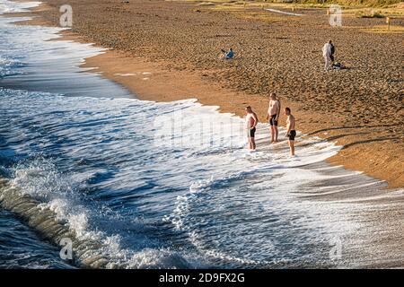 Vita quotidiana. Gruppo di uomini che guardano le onde sulla spiaggia in una giornata soleggiata ma fredda. Foto Stock