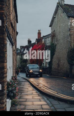 Frome, Regno Unito - 04 ottobre 2020: Auto con fari sulla guida oltre case di pietra coperte di fogliame colorato su una strada a Frome, Somerset, Regno Unito. Foto Stock