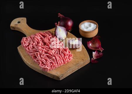 La carne tritata e le cipolle rosse giacciono su un tagliere di legno. Sale shaker con sale. Sfondo nero Foto Stock