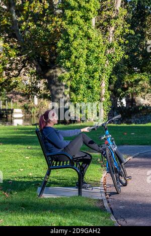 una donna seduta su una panchina con una bicicletta accanto a lei sotto un bell'albero in un parco durante la stagione autunnale o autunnale. Foto Stock