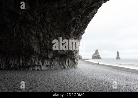 Vista incredibile sulla spiaggia nera e le scogliere di Troll Toes in tempo nuvoloso. Solitario turista con treppiede che va sulla costa. Reynisdrangar, Vik, Islanda. Fotografia di paesaggio Foto Stock