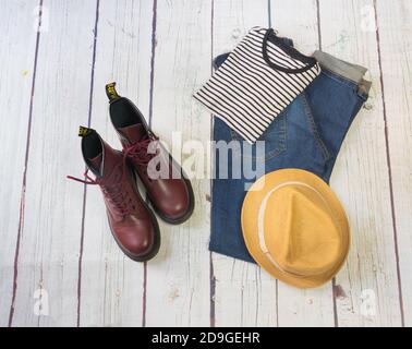 Ancora vita con stivali militari borgogna, jeans, t-shirt a righe e cappello, su uno sfondo di legno bianco Foto Stock