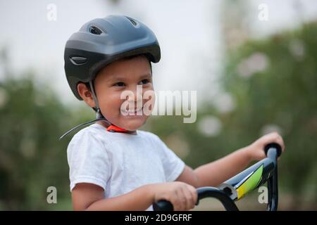 Un ragazzo di quattro anni sorride, mentre indossa il casco per bicicletta per sicurezza e si tiene sulle barre dell'impugnatura della bicicletta. Foto Stock