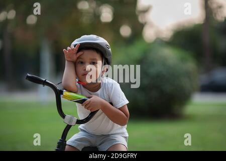 Un ragazzino che sembra sfinito o mostra la difficoltà a guidare una bicicletta per la prima volta tenendo la mano sulla fronte e guardando infelice. Foto Stock