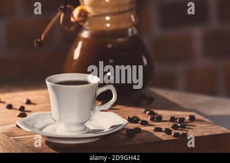 Tazza di caffè esagonale bianca con chicchi di caffè arabica rotondi prodotti in Brasile. Macchina per il caffè sfocata sullo sfondo. Paesaggio rustico. Foto Stock