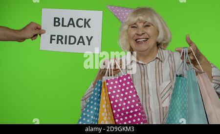 L'iscrizione pubblicitaria Black Friday appare accanto alla nonna gioiosa. Donna anziana con borse per lo shopping, balli, festeggiamenti, in cerca di soddisfatti con prezzi bassi per lo shopping, gioendo sconti Foto Stock