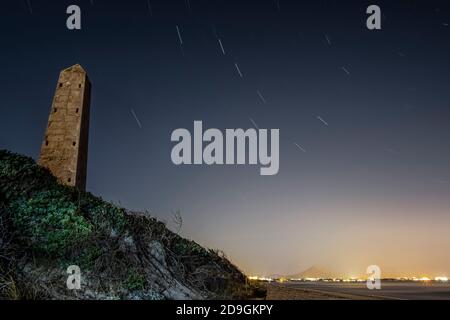 Immagine notturna di una spiaggia con un monolito di pietra, luci e città sullo sfondo e una linea di una stella che forma una circumpolare, a Playa de Muro, Palma de Foto Stock