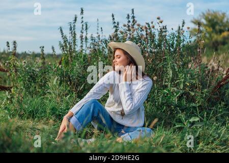 Giovane bella donna bruna vestita di un maglione bianco, jeans e cowboy cappello di paglia seduta su erba verde, sorridente e ridente durante il tramonto Foto Stock