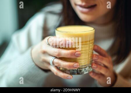 Giovane caucasica sorridente e tenente un bicchiere di caldo latte curcuma sul tavolo Foto Stock