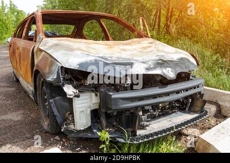 Vista frontale di un'auto arrugginita bruciata durante il giorno d'estate, paraurti mancante, cofano sgualcito. Foto Stock