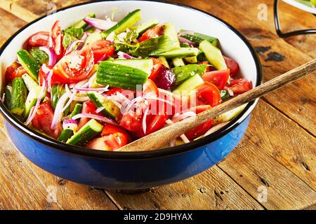 Insalata di cetrioli, pomodori, cipolle ed erbe in una ciotola blu-bianca su fondo di legno. Foto Stock