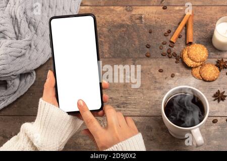 Immagine mockup delle mani di donna che tengono il telefono cellulare con schermo bianco da scrivania mentre si beve caffè in caffetteria. La donna a un tavolo in un caffè usa un mmo Foto Stock