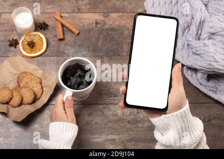 Telefono cellulare mock-up. Donna a un tavolo rustico in legno beve caffè e guarda un telefono cellulare mock-up in inverno. Vista dall'alto Foto Stock